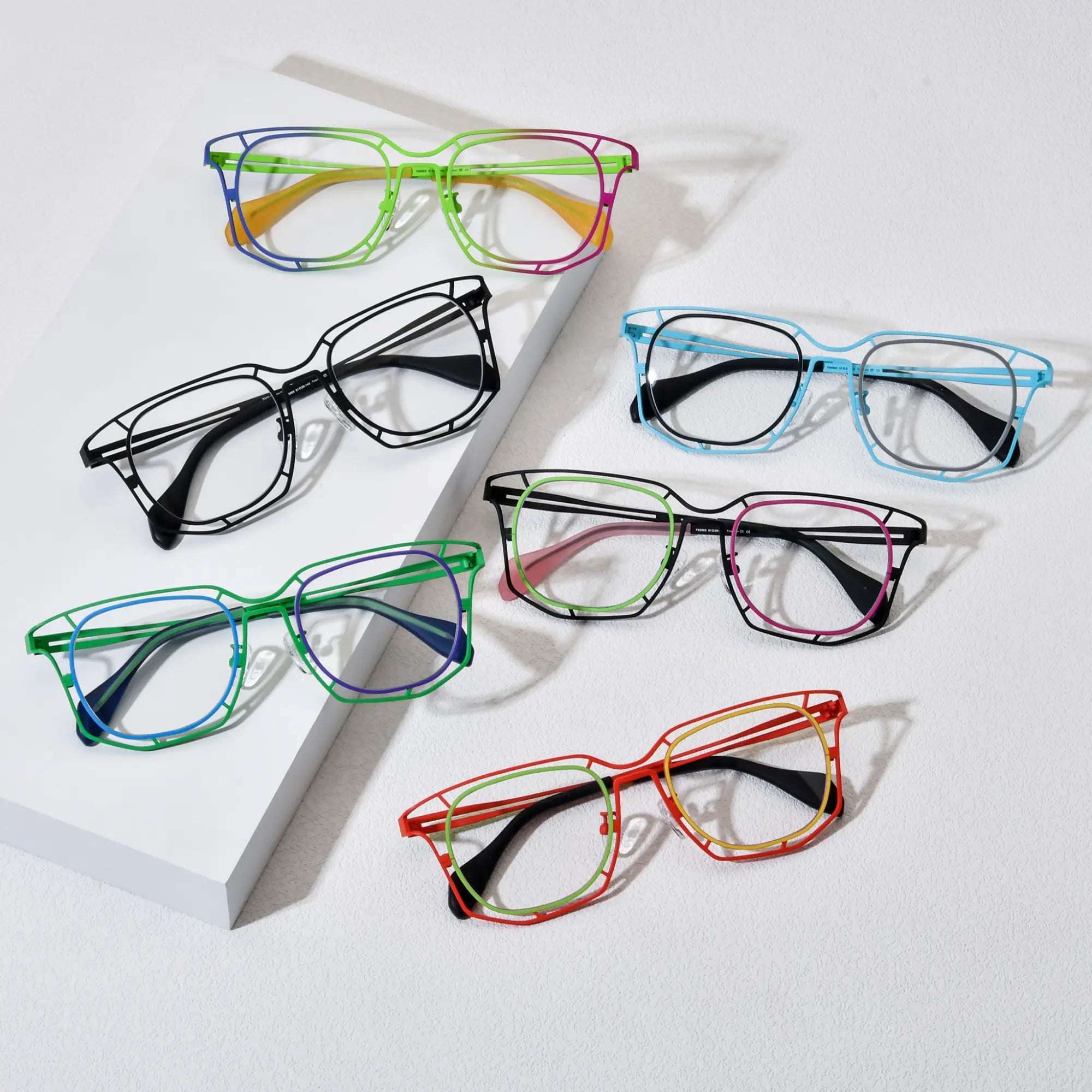 Display of VoyueLens™ VL185865 eyeglasses in six color variants – Rainbow Burst, Sky Blue, Graphite Black, Mint Pink Fusion, Emerald Twilight, and Amber Blaze. Polygonal titanium frames arranged on a textured background.